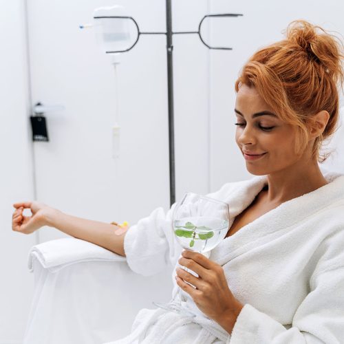 Waist up portrait view of the charming woman in white bathrobe sitting in armchair and receiving IV infusion. She is holding glass of lemon beverage and smiling